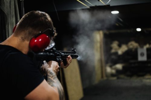 mężczyzna z tatuażami w czerwonych słuchawkach strzeleckich celujący z karabinu na strzelnicy