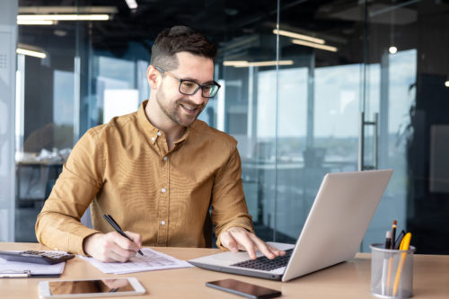 uśmiechnięty mężczyzna w okularach siedzi przy biurku w biurowej przestrzeni i pracuje na laptopie oraz z dokumentami