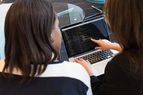 dwie kobiety patrzące na ekran laptopa na którym widać kod źródłowy, kobieta wskazuje palcem na ekran