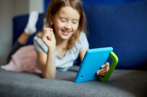 Dziewczynka leżąca na kanapie, Dziecko uzależnione od technologii, lubiące grać w gry online na cyfrowym tablecie, korzystać z aplikacji, przeglądać strony internetowe w domu