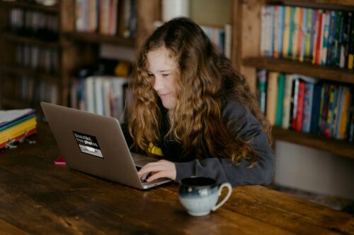 nastolatka z kręconymi włosami i lekkim uśmiechem na twarzy siedząca przy stole w bibliotece pracuje na laptopie