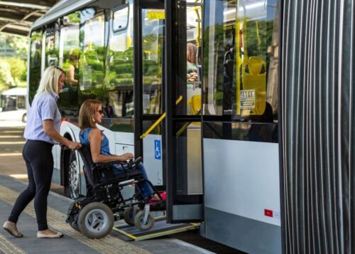 Obraz przedstawia kobietę na wózku inwalidzkim wjeżdżającą do autobusu za pomocą rampy dla niepełnosprawnych. Wózek jest nowoczesny, elektryczny. Kobieta jest ubrana w dżinsy i niebieską koszulkę bez rękawów. W tle widać wnętrze autobusu z jasnymi, żółtymi poręczami i miejscami siedzącymi. Przy kobiecie stoi inna kobieta, która pomaga jej wejść na rampę, popychając wózek. Ona ma na sobie jasną bluzkę i ciemne spodnie. Autobus jest biały z czarnymi oknami, a na drzwiach widoczny jest niebieski symbol wskazujący na dostępność dla osób niepełnosprawnych. Scena rozgrywa się na przystanku autobusowym, a w tle można dostrzec drzewa i inne autobusy. Atmosfera na obrazie jest spokojna i pokazuje przyjazne środowisko dla osób niepełnosprawnych.